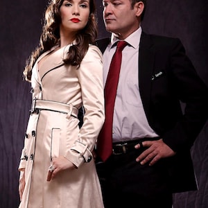 May include: A woman wearing a white trench coat with black trim stands next to a man in a black suit and red tie. The man has a name tag that says "Lynch".