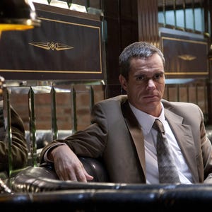 May include: A man in a brown suit sits in a leather armchair. He is wearing a white shirt and a patterned tie. The man has a serious expression on his face.