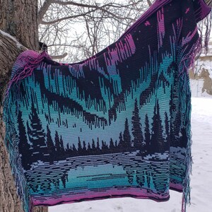 May include: A crocheted blanket with a colorful aurora borealis design and a fringe border. The blanket is hanging on a tree branch in a snowy landscape.