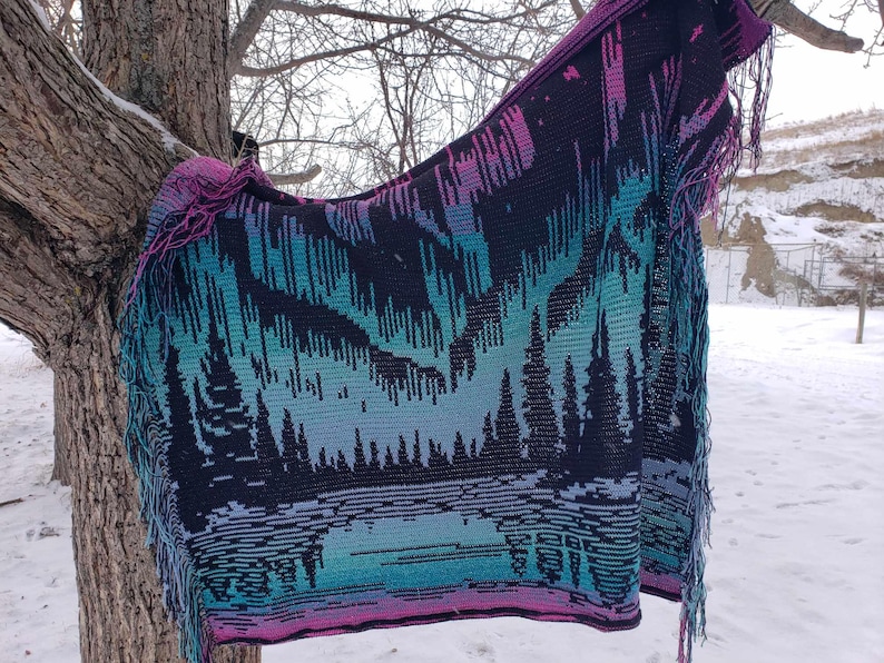 May include: A crocheted blanket with a colorful aurora borealis design and a fringe border. The blanket is hanging on a tree branch in a snowy landscape.