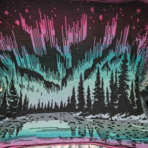 May include: A crocheted throw blanket with a black background and a colorful aurora borealis design. The aurora is depicted in shades of pink, purple, blue, and green. The bottom of the blanket features a dark forest silhouette.