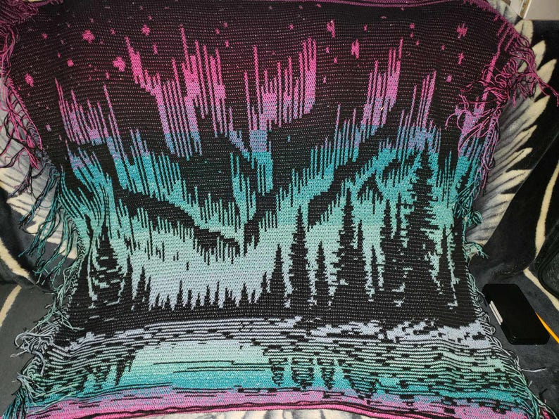 May include: A crocheted throw blanket with a black background and a colorful aurora borealis design. The aurora is depicted in shades of pink, purple, blue, and green. The bottom of the blanket features a dark forest silhouette.