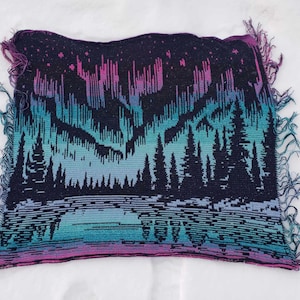 May include: A woven tapestry featuring a dark blue background with a colorful aurora borealis scene. The aurora is depicted in shades of pink, purple, blue, and green. A row of dark green trees are silhouetted in front of the aurora.