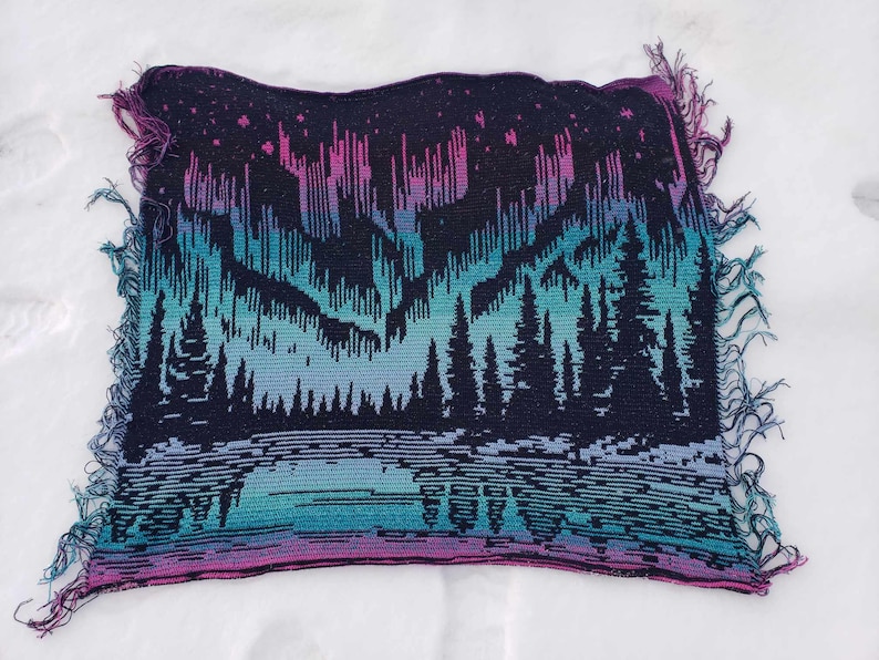 May include: A woven tapestry featuring a dark blue background with a colorful aurora borealis scene. The aurora is depicted in shades of pink, purple, blue, and green. A row of dark green trees are silhouetted in front of the aurora.