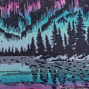 May include: A woven tapestry with a fringed edge features a dark blue background with a colorful aurora borealis design in shades of pink, purple, blue, and green. The aurora borealis is depicted above a dark forest silhouette.