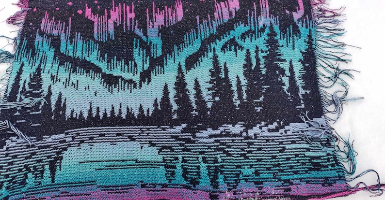 May include: A woven tapestry with a fringed edge features a dark blue background with a colorful aurora borealis design in shades of pink, purple, blue, and green. The aurora borealis is depicted above a dark forest silhouette.