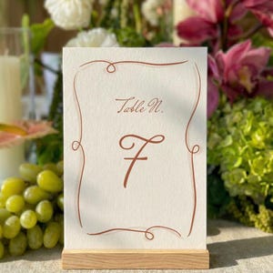 Può includere: Un segnaposto rettangolare con il numero 7 in carattere marrone. La carta ha un bordo decorativo marrone e le parole "Table N." in alto. È posizionato su una base di legno, adatto per matrimoni o eventi.