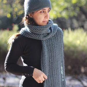 May include: A knitted hat and scarf set in a muted teal color. The scarf is long and chunky, wrapped around the neck. The hat is a simple beanie style. The person is wearing a black long-sleeved shirt.