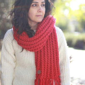 Może przedstawiać: Kobieta w białym swetrze i czerwonym dzianym szaliku z drewnianym guzikiem i długimi frędzlami. Szalik jest owinięty wokół szyi, a frędzle sięgają jej klatki piersiowej.