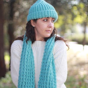 May include: A knitted turquoise beanie and scarf set. The beanie is a close-fitting style, and the scarf is long and chunky. The person is wearing a white sweater. The background is blurred with natural light.