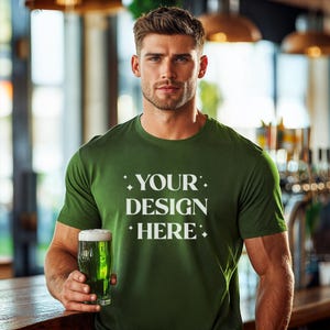 Puede incluir: Un hombre con una camiseta verde con el texto "YOUR DESIGN HERE" impreso en la parte delantera. Está sosteniendo un vaso de cerveza verde.