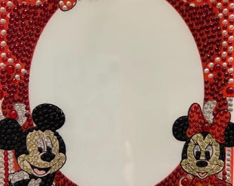 May include: A red and white photo frame decorated with red and silver rhinestones. Mickey and Minnie Mouse are featured on the frame. The frame has a white oval opening.