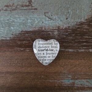 May include: A heart-shaped pendant made from a dictionary page, featuring the word "traveler." The pendant is set against a rustic, multi-toned wooden surface, creating a vintage aesthetic. The text is clear and legible.