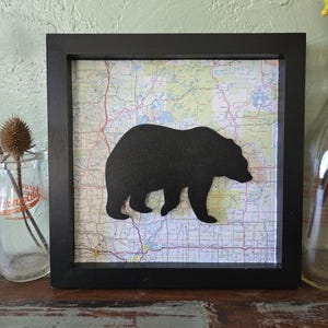 May include: A black bear silhouette is mounted on a vintage map in a black frame. The map is a detailed road map with a light green, yellow, and brown color scheme. The frame is on a wooden surface.