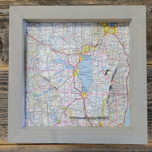 May include: A framed vintage map of Wisconsin with the state outline cut out. The map is in shades of blue, green, and brown. The frame is a light gray wood.