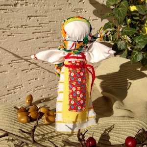 May include: A traditional folk doll with a white dress, yellow and red striped skirt, and a colorful headscarf. The doll is standing on a beige fabric background with brown branches and red berries.