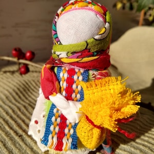 May include: A colorful handmade rag doll with a yellow fabric bundle in its arms. The doll is wearing a red, green, blue, and white patterned dress and a pink and green patterned head scarf.