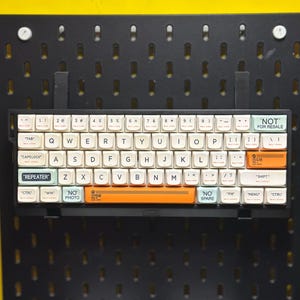 May include: A compact, black mechanical keyboard with white keycaps and orange and green accent keys. The keyboard is mounted on a black pegboard. The keycaps feature English characters and special function labels.