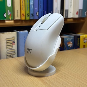 Può includere: Un mouse per computer bianco con un supporto abbinato. Il mouse ha un design elegante con una rotella di scorrimento e pulsanti. Il supporto è progettato per tenere il mouse in posizione verticale. Il mouse ha un logo sul lato.