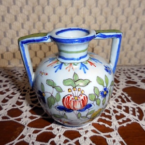 May include: A small, hand-painted ceramic vase with two handles. The vase is white with a floral design in red, blue, green, and yellow. The handles and rim are accented with blue and green paint.