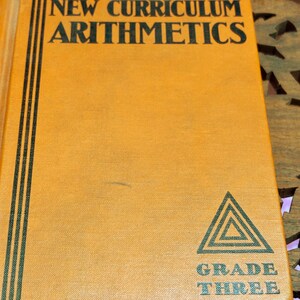 May include: A hardcover book with a bright orange cover featuring black text that reads The NEW CURRICULUM ARITHMETICS. The cover includes three vertical black lines on the left side and a black triangle logo above the text GRADE THREE.