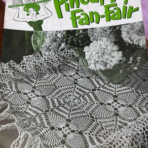 May include: Vintage "Pineapple Fan-Fair" crochet pattern book cover. The cover features a green and white illustration of a woman holding pineapples, with the title in large green letters. A white crocheted doily is in the foreground.