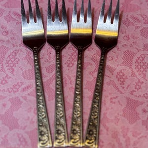 May include: Four silver forks with tines and ornate floral designs on the handles. The forks are arranged vertically against a pink lace background, creating a vintage aesthetic. The forks are made of metal.