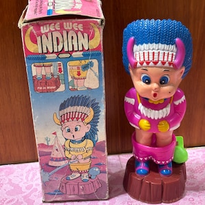 May include: A vintage toy featuring a Native American figure with blue hair, a pink outfit, and a red base. The toy is accompanied by its original box, which has the words "WEE WEE INDIAN" printed on it.