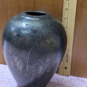 May include: A dark, speckled vase with a wide opening. The vase has a rounded body and a slightly textured surface. The color is primarily black with lighter, speckled accents. A wooden ruler is visible in the background.