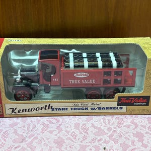 May include: Die-cast metal 1925 Kenworth stake truck with barrels. The red truck has black accents and the text "True Value" on the side. The truck is in its original packaging.
