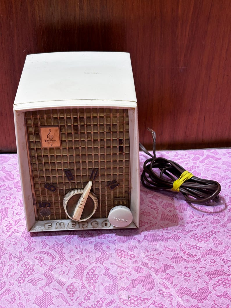 May include: Vintage Emerson radio with a cream-colored cabinet and a brown speaker grill. The radio features a tuning dial and control knobs. The word "Emerson" is printed on the front. A brown power cord is attached.