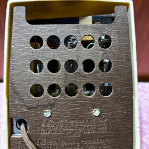May include: Close-up of the back of a vintage Emerson radio. The cream-colored plastic casing frames a brown, perforated panel with several visible tubes. Text on the panel reads "EMERSON RADIO & PHONOGRAPH CORPORATION NEW YORK U.S.A."