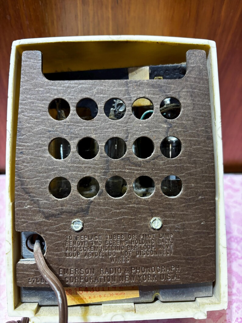 May include: Close-up of the back of a vintage Emerson radio. The cream-colored plastic casing frames a brown, perforated panel with several visible tubes. Text on the panel reads "EMERSON RADIO & PHONOGRAPH CORPORATION NEW YORK U.S.A."