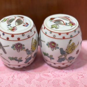 May include: Two white ceramic jars with lids. Each jar is decorated with colorful floral and insect designs. The lids have a handle with two openings. The jars are on a pink lace surface.