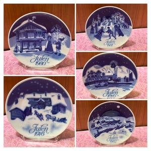 May include: A collection of five circular porcelain plates featuring blue and white winter scenes. Each plate displays a unique snowy landscape, figures, or buildings, with the text Julen followed by the years 1978, 1980, 1981, 1982, and 1979.