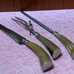 May include: A set of vintage carving tools, including a knife, fork, and sharpening steel. The handles are a light brown color with a white base. The tools are arranged on a pink lace tablecloth.