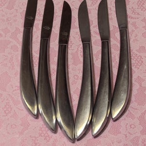 May include: Six stainless steel butter knives arranged vertically. Each knife has a curved handle and a rounded blade. The knives are displayed on a pink floral patterned surface.