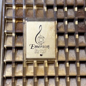 May include: Close-up of a vintage radio or television grill with a gold-toned metal finish. The grill has a grid pattern with a rectangular Emerson logo in the center. The logo includes the words "Emerson Television and Radio" in a classic font.