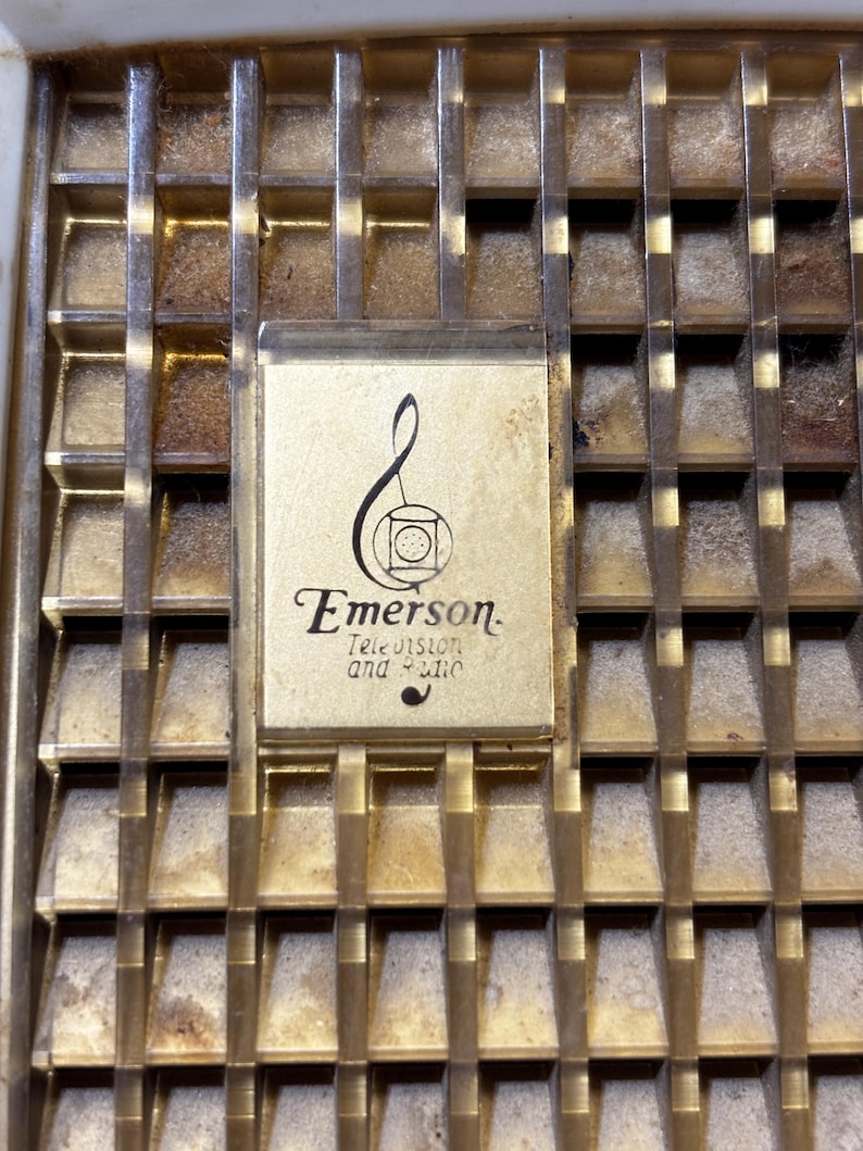 May include: Close-up of a vintage radio or television grill with a gold-toned metal finish. The grill has a grid pattern with a rectangular Emerson logo in the center. The logo includes the words "Emerson Television and Radio" in a classic font.
