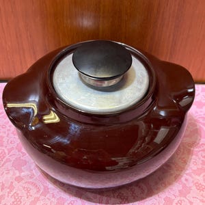 May include: A round, dark brown ceramic pot with a glossy finish and two integrated side handles. The pot features a removable circular lid with a brushed silver-tone metal center and a small, smooth black knob handle centered on top.