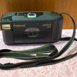 May include: A black Polaroid Captiva SLR camera with a flash and a lens. The camera has a black and green strap. The camera has the words "Polaroid" and "Captiva SLR" printed on it.