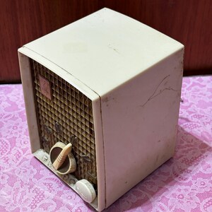 May include: Vintage off-white radio with a brown speaker grill and two cream-colored knobs. The radio has a boxy shape and a small emblem on the speaker. The radio is sitting on a pink floral patterned surface.