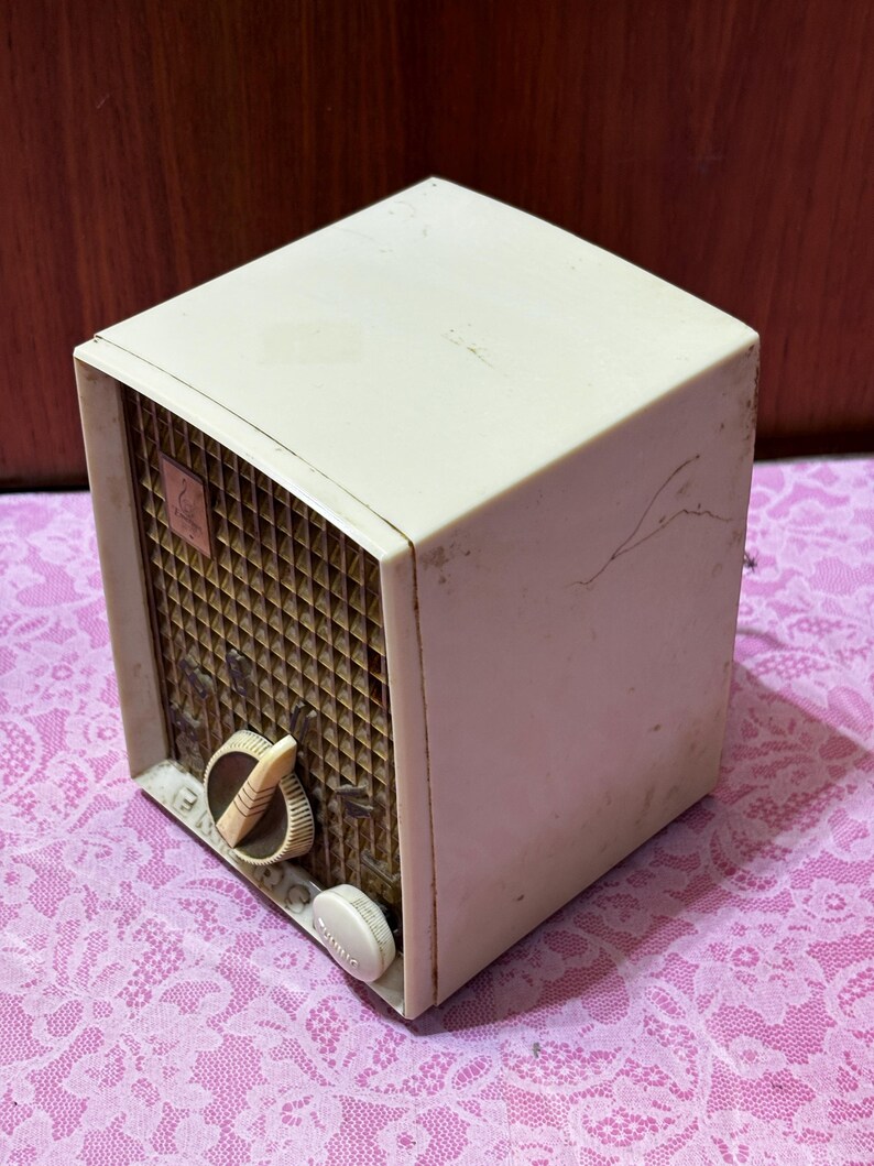 May include: Vintage off-white radio with a brown speaker grill and two cream-colored knobs. The radio has a boxy shape and a small emblem on the speaker. The radio is sitting on a pink floral patterned surface.