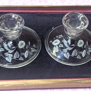 May include: Two clear glass candle holders with a floral and bird design etched on the base. The design features flowers, leaves, and a bird in flight. The candle holders are round with a small cup to hold the candle.