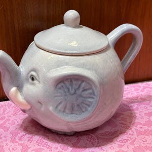 May include: A light blue ceramic teapot shaped like an elephant with a removable lid. The teapot features a glossy, crackled glaze finish, a curved handle, a spout, and molded ear and tusk details on the front of the rounded body.
