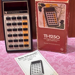 May include: A vintage Texas Instruments TI-1250 electronic calculator with its original box and manual. The calculator is black with orange and white keys. The box is red with a picture of the calculator. The manual is white with black text.