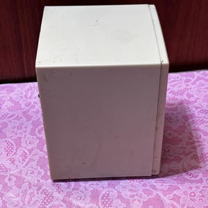 May include: A rectangular, off-white box with a slightly textured surface. The box is sitting on a pink lace patterned surface. The box appears to be made of plastic or a similar material.