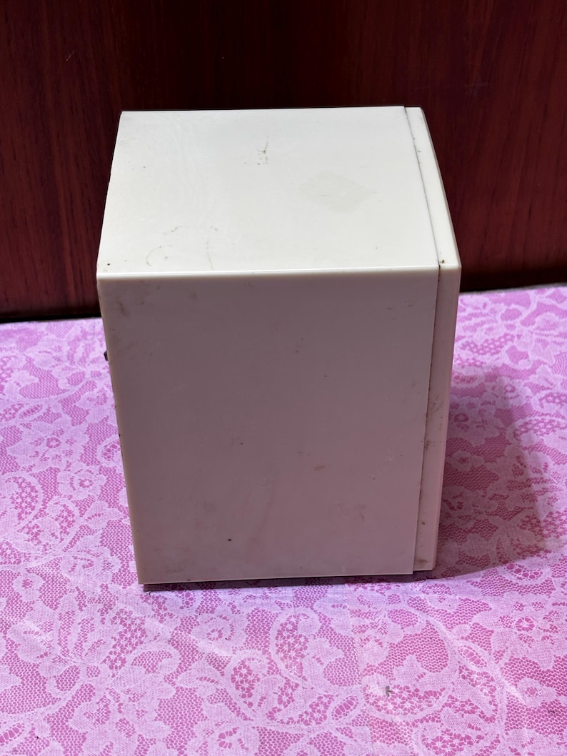 May include: A rectangular, off-white box with a slightly textured surface. The box is sitting on a pink lace patterned surface. The box appears to be made of plastic or a similar material.