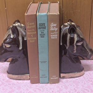 May include: A set of three brown bookends shaped like horse heads. The bookends are holding up three books with the title "Gene Autry" on the spines. The books are titled "Badmen of Broken Bow", "Big Valley Chad", and "Redwood Pirates".