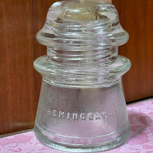 May include: Clear glass Hemingray-17 insulator. The vintage electrical insulator has a rounded top and base with a cylindrical body. The name "HEMINGRAY-17" is embossed on the side. The insulator is sitting on a pink lace tablecloth.
